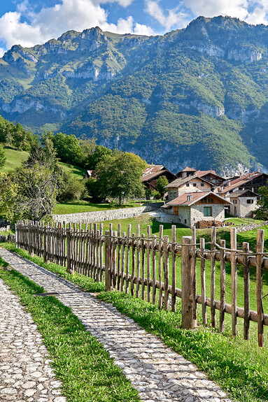 It is a clear, sunny day. A stone path surrounded by a bright green meadow and flanked by a wooden fence leads us to Irone. Irone is a ghost village: in 1630 it was completely depopulated because of the plague. Now in summer it repopulates, in fact the houses in the picture are neat and well kept. In the background is Mount Iron and a deep blue sky with a few white clouds. 