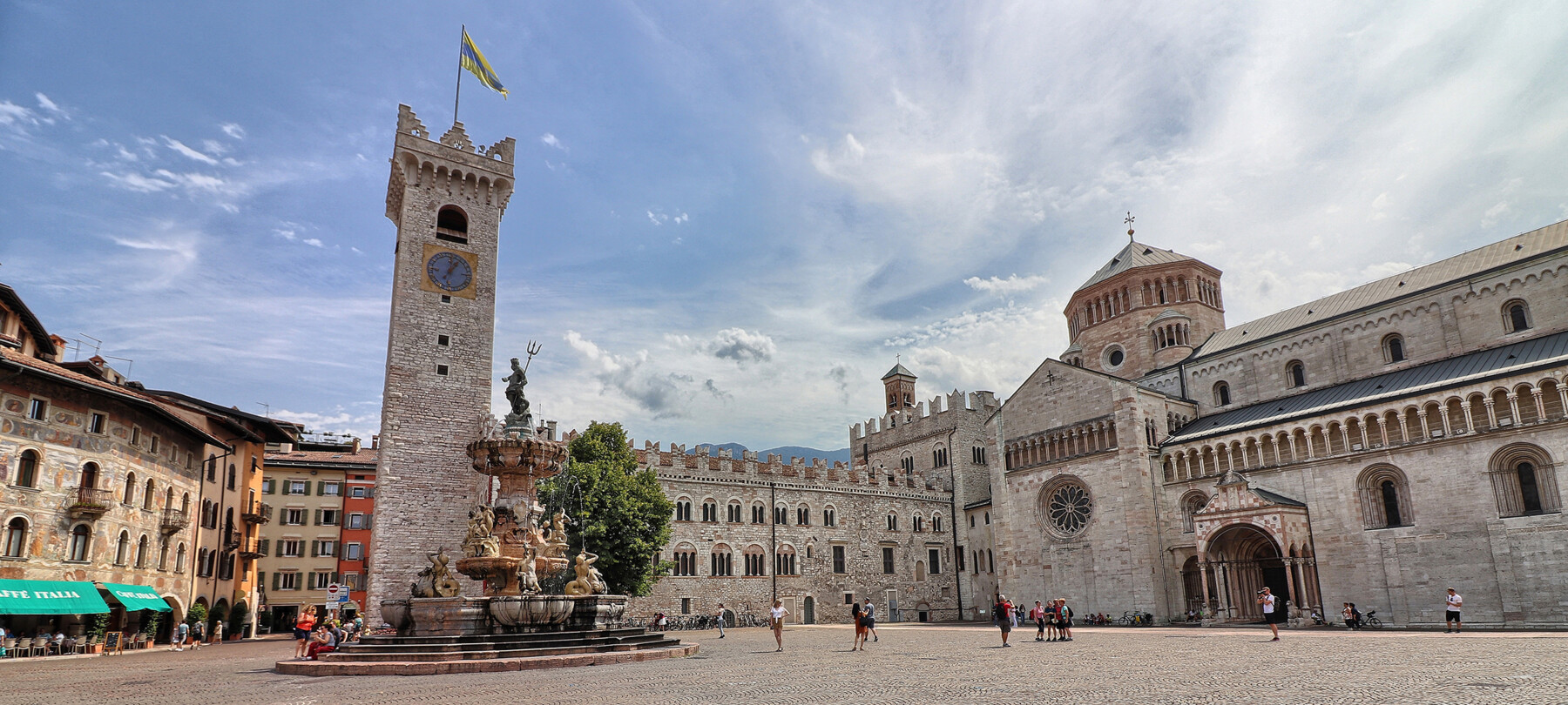 Piazza Duomo in Trient. Im Hintergrund der Stadtturm, der Palazzo Pretorio und die Kathedrale San Vigilio, die Kathedrale der Stadt. In der Mitte: der Neptunbrunnen. Hinter dem Brunnen steht der einzige Baum auf dem Platz: eine Linde. Der Platz ist auf dem Bild belebt. Einige Leute sitzen auf den Stufen des Brunnens, jemand macht Fotos und eine kleine Gruppe posiert vor der Kathedrale, um sich fotografieren zu lassen. Alle tragen Sommerkleidung. Der blaue Himmel ist mit dem Weiß leichter Wolken durchzogen.