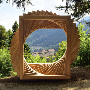Bosco Arte Stenico, ein Werk von Maurizio Corradi. Das Werk, das sich an einem Panoramapunkt des Weges befindet, ist eine große Spirale aus Holzelementen, die wie ein offenes Fenster auf das Panorama wirkt. In der Mitte, in der Ferne, befindet sich die Burg von Stenico. Um das Werk herum die Bäume des Waldes.