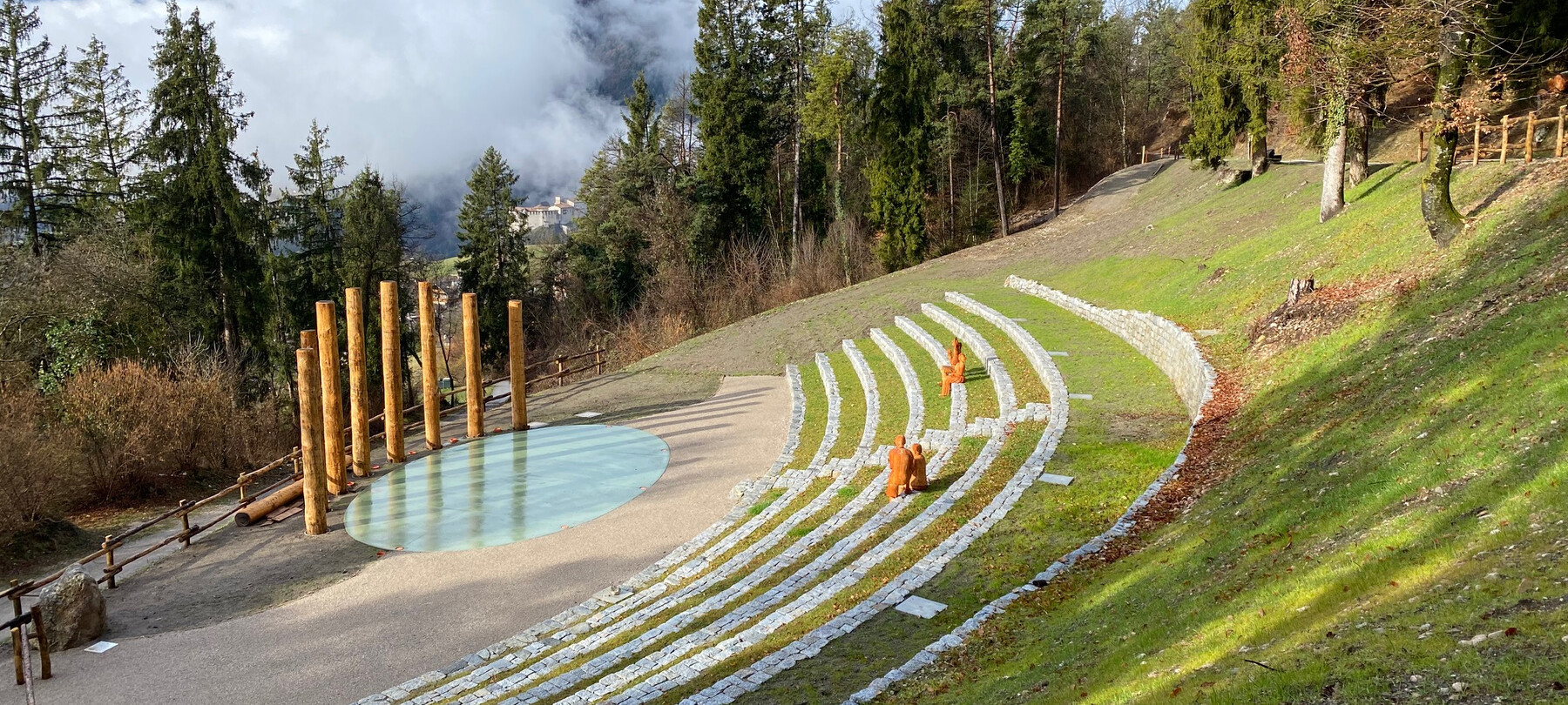 Installazione artistica di Bosco Arte Stenico. Un anfiteatro naturale, nel quale gradini di pietra seguono la pendenza della montagna. Sui gradini ci sono quattro figure umane, sedute a due a due. Sono statue in legno, rivolte verso l’arena dell’anfiteatro, dove si trovano uno specchio d’acqua perfettamente ovale e, dietro, un ventaglio di alte colonne in legno. Tutto intorno, pini, larici e faggi. In lontananza, nuvole bianche e grigie riempiono il cielo e coprono la montagna, che fa capolino solo ogni tanto dove le nuvole si aprono.