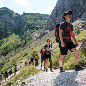 Mehrere Personen wandern hintereinander auf einem Pfad in den Brenta-Dolomiten. Die zweite Person in der Reihe – eine Frau mit einer Prothese am linken Bein– wandert mithilfe zweier Trekkingstöcke, wie auch andere Personen in der Gruppe.