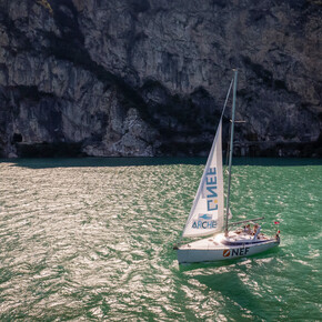 Una barca a vela della Cooperativa Archè attraversa le acque verdi e illuminate dal sole del lago di Garda. Alle spalle, una parete rocciosa in ombra chiude la scena come un sipario. | © ©Trentino Marketing_Gianluca Prati_Accessibilita_Riva del Garda_2024_00001
