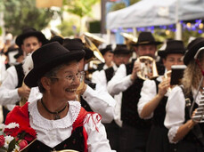 The photograph portrays a group of musicians in traditional costume, likely during a folk festival. In the foreground, an elderly woman smiles, wearing a red and white dress with a black hat, holding a golden horn and a bouquet of red and white flowers. In the background, other musicians in similar attire play instruments and wear dark hats. The atmosphere is festive and cheerful, with lights and decorations suggesting an outdoor event in a square or street. The image conveys a strong sense of tradition, community, and shared joy.