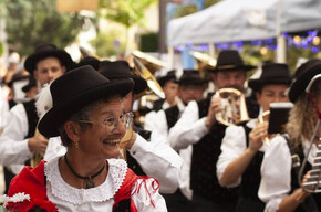 The photograph portrays a group of musicians in traditional costume, likely during a folk festival. In the foreground, an elderly woman smiles, wearing a red and white dress with a black hat, holding a golden horn and a bouquet of red and white flowers. In the background, other musicians in similar attire play instruments and wear dark hats. The atmosphere is festive and cheerful, with lights and decorations suggesting an outdoor event in a square or street. The image conveys a strong sense of tradition, community, and shared joy.