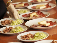 A wooden table with several neatly arranged white plates. Each plate features a combination of cold cuts, cheese, beans, and chopped cabbage salad. A hand wearing a white glove delicately arranges the plates, suggesting a professional setup for a catering service.