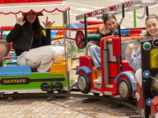 The image shows a red toy train with four children sitting on it, having fun. The train is working and moving along small tracks in the town square.