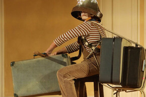 L'immagine mostra la protagonista durante lo spettacolo mentre è in partenza con le sue valigie, un imbuto in testa e delle lucine sopra