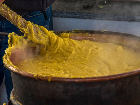 La foto cattura il primo piano di un paiolo colmo di polenta mentre un uomo mescola con un grande bastone in legno