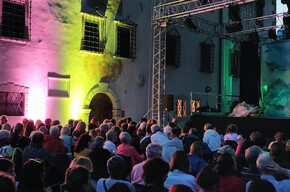L'immagine mostra un pubblico di circa 200 persone sedute che aspettano lo spettacolo. Le sedie sono poste nel cortile di un palazzo ottocentesto illuminato da luci gialle e rosa. Sul palco ci sono delle strutture probabilmente utili allo spettacolo che sta per iniziare. Le persone sono sorridenti e non vedono l'ora che inizi. 