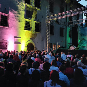L'immagine mostra un pubblico di circa 200 persone sedute che aspettano lo spettacolo. Le sedie sono poste nel cortile di un palazzo ottocentesto illuminato da luci gialle e rosa. Sul palco ci sono delle strutture probabilmente utili allo spettacolo che sta per iniziare. Le persone sono sorridenti e non vedono l'ora che inizi. 