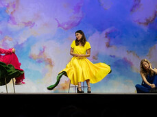 On the stage, there are three women. The one in the middle is sitting on a chair and wearing a yellow dress. She is talking to the woman on the left, who is also seated on a flower-shaped chair and dressed in red. On the other side, a woman is sitting on the floor, gently touching a large artificial flower.