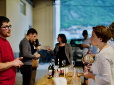 In the foreground, the young man on the left is explaining to the woman with short hair the products from his company, with which he has prepared a platter of cured meats. The woman is drinking a glass of red wine. In the background, the rest of the group is finishing the winery tour and beginning the tasting.