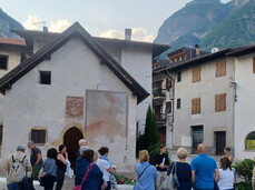 A group of about 20 people is visiting the small church of Roveré della Luna together with a historical guide. It's evening, but not yet dark. At the moment, they are outside the church, admiring a fresco.