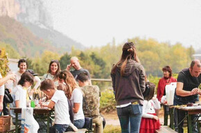 A group of children and adults take part in an outdoor activity surrounded by nature, with craft tables, games, and a warm, communal atmosphere. The event feels friendly and relaxed, ideal for a local gathering. Hills and vineyards can be seen in the background.