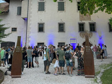 A group of people gathers in front of a historic building with walls illuminated by blue lights. The atmosphere is friendly and sociable; it is a cultural and food-and-wine event. Two metal structures bearing the word "MEZZOCORONA" mark the entrance.