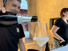 In the foreground, a person is serving a glass of Trento Doc sparkling wine to someone, carefully pouring the wine. Next to him, along the bar counter, there's a young woman at the register taking orders.