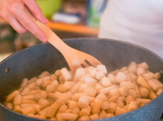 The image shows a person cooking in a pan filled with small pieces of white asparagus, which appear to be sautéed or stewed. The person is stirring the asparagus with a wooden spoon, a utensil often used to avoid damaging the surface of the pan. Steam is rising from the pan, indicating that the food is cooking and probably close to being ready. The background is blurred but reveals a warm and welcoming environment, with warm colors and details suggesting a kitchen or a culinary workshop.