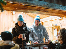 Two local young people, a girl and a boy, are manning the little food stall at the Christmas market. They are selling mugs of mulled wine and hot chocolate to other guys.