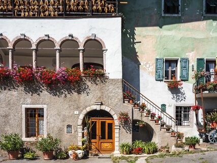 A view of San Lorenzo in Banale on a sunny day. The red and purple spots of the many pots of flowers, perhaps geraniums, stand out in the picture: they are hanging from the windows and the edge of a terrace surrounded by arches, leaning on the steps and on a small balcony. The sunshine and these bright colours convey a sense of cheerfulness.